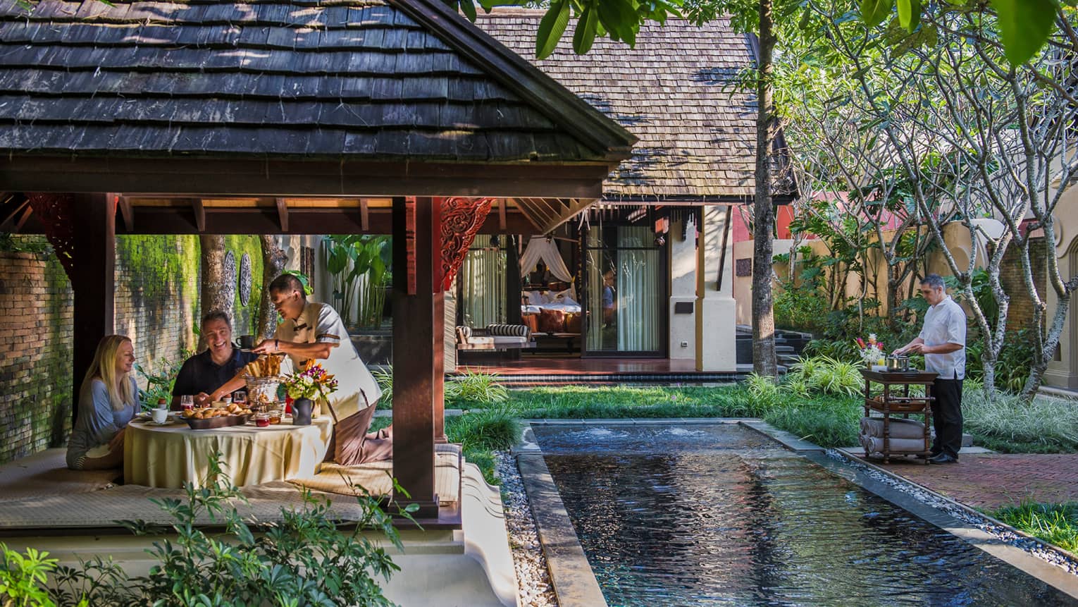 Staff serve couple under covered dining gazebo by long villa pool