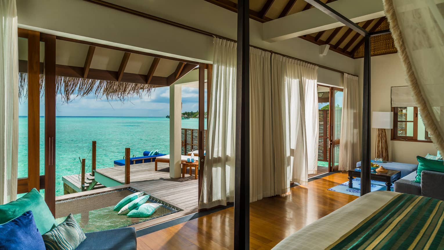 Sunrise Water Villa blue chair with pillows by poster bed across from open wall, patio, ocean views
