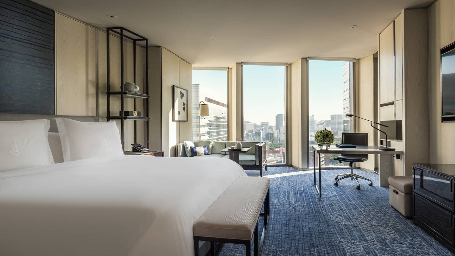 Deluxe Room hotel bed with bench, small sofa and table by floor-to-ceiling windows