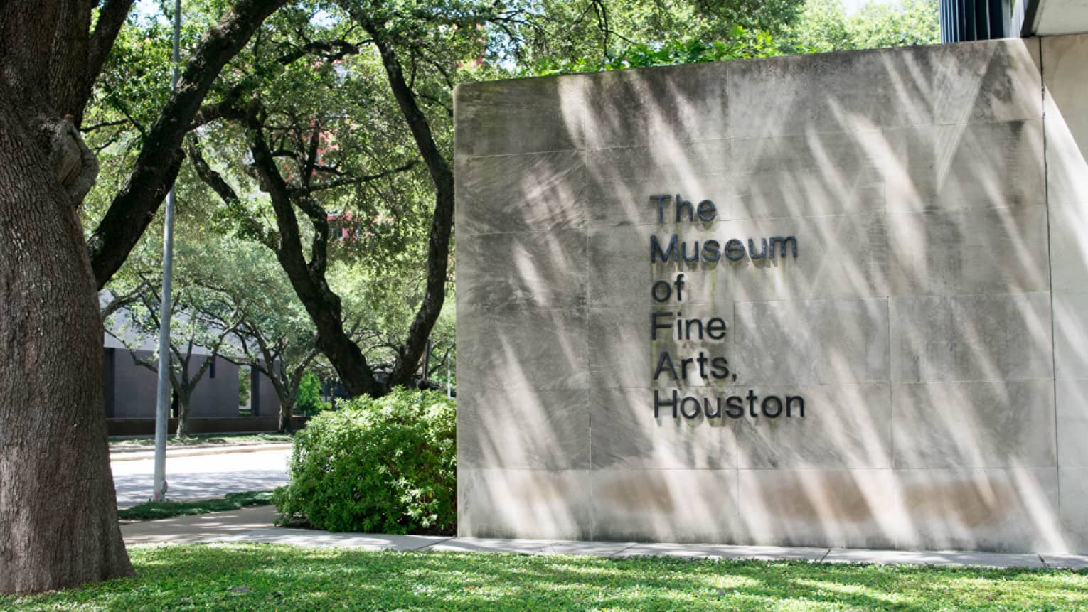 The Museum of Fine Arts Houston sign on stone wall by green lawn, trees
