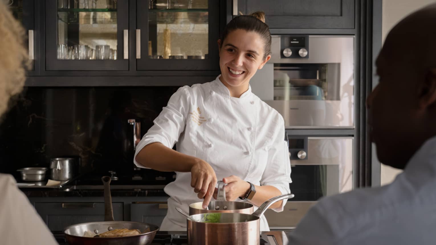 Smiling female chef stirs food in a copper pot as two other people look on