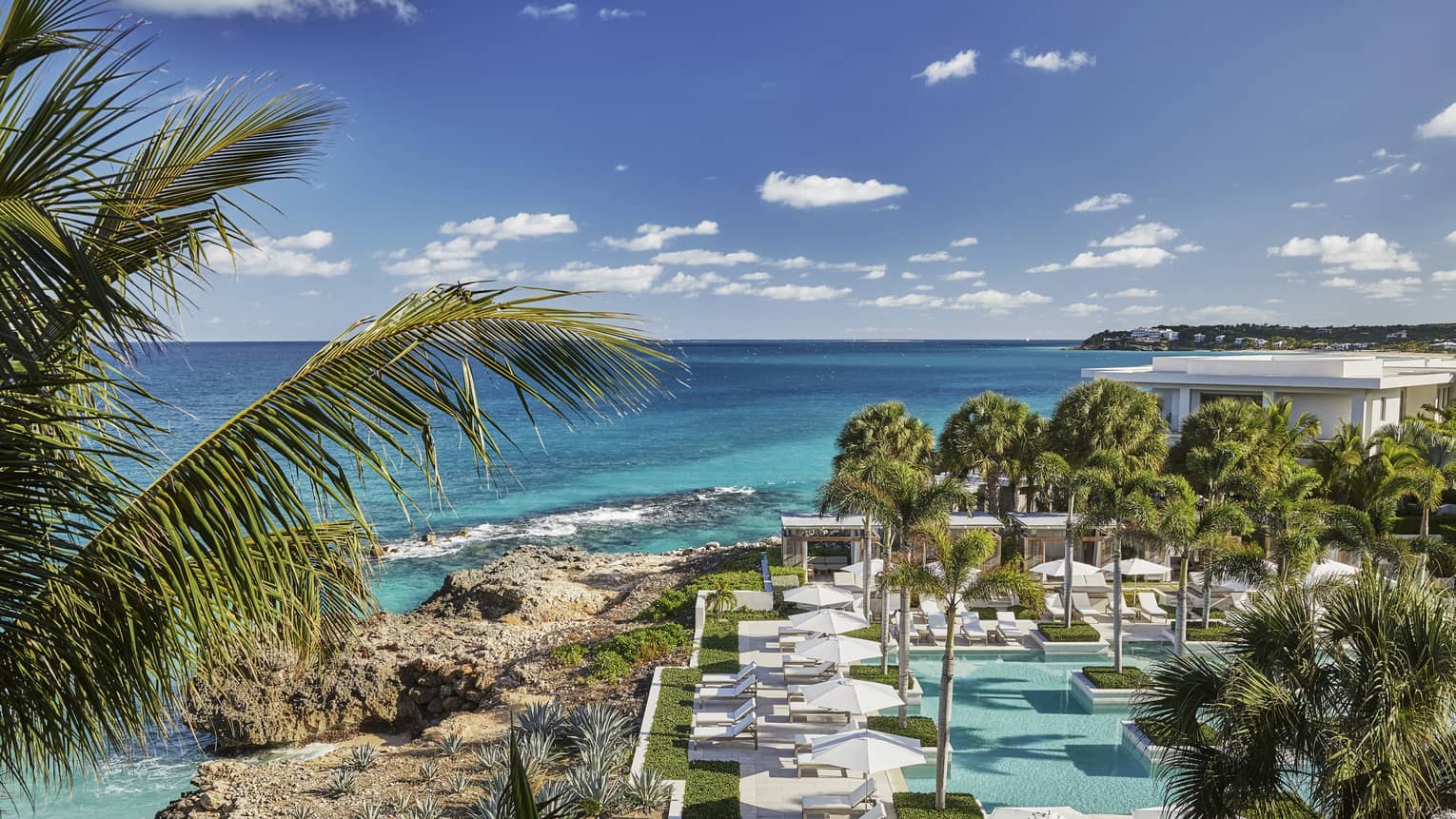 Looking down over Anguilla outdoor pool, patio, large rocks along blue-green ocean waterfront, blue sky