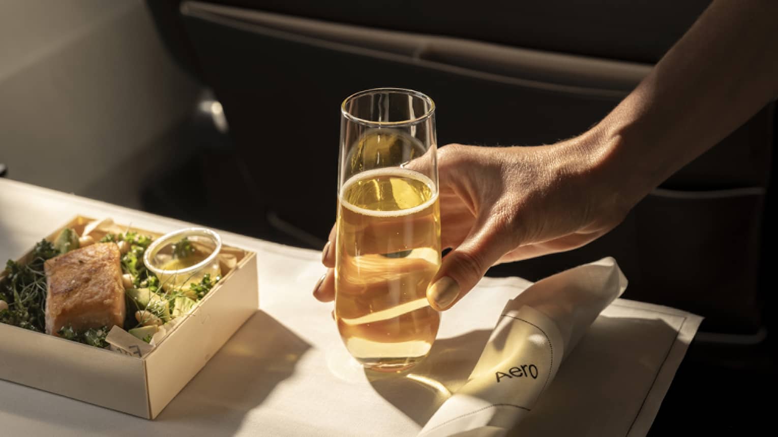 A person's hand sets down a stemless champagne flute next to a salad on an airplane tray table