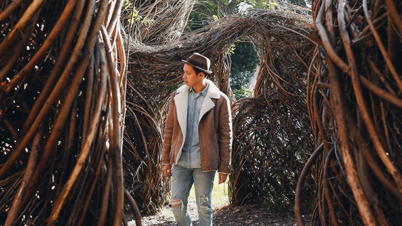 Man in shearling coat, fedora hat walks through twisted branches, trees