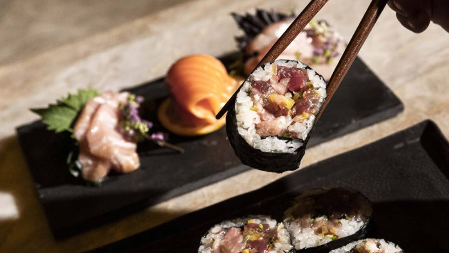 Two plates of sushi rolls and cuts of fish. A hand holds a piece of a roll in chopsticks.