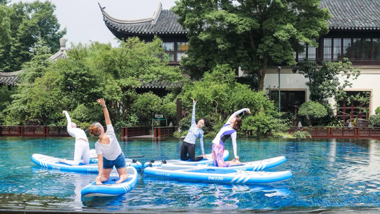 Four people kneel on paddleboards arranged like petals in a tranquil pool, one arm stretched skyward, amid lush foliage.