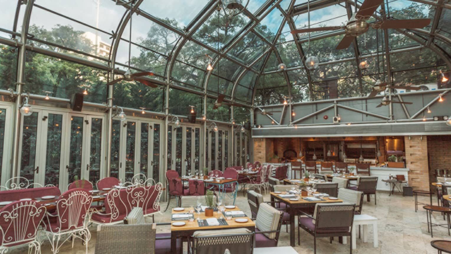 Tables are arranged throughout an enclosed outdoor patio with glass walls and ceilings