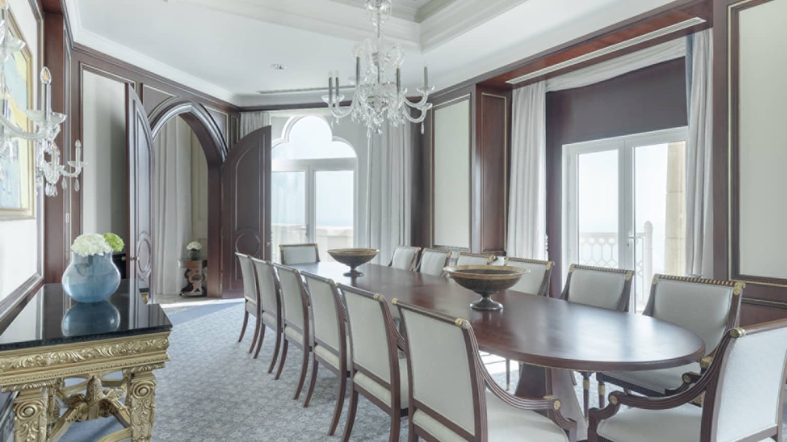 Elegant dining room with 14 seats, long wooden table, natural light, chandelier, ornate golden side table
