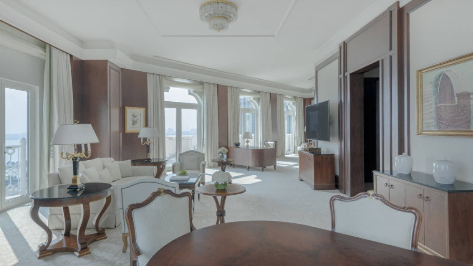 Large living area of hotel suite, with dark wooden dining table, white sofa and many windows lining the wall