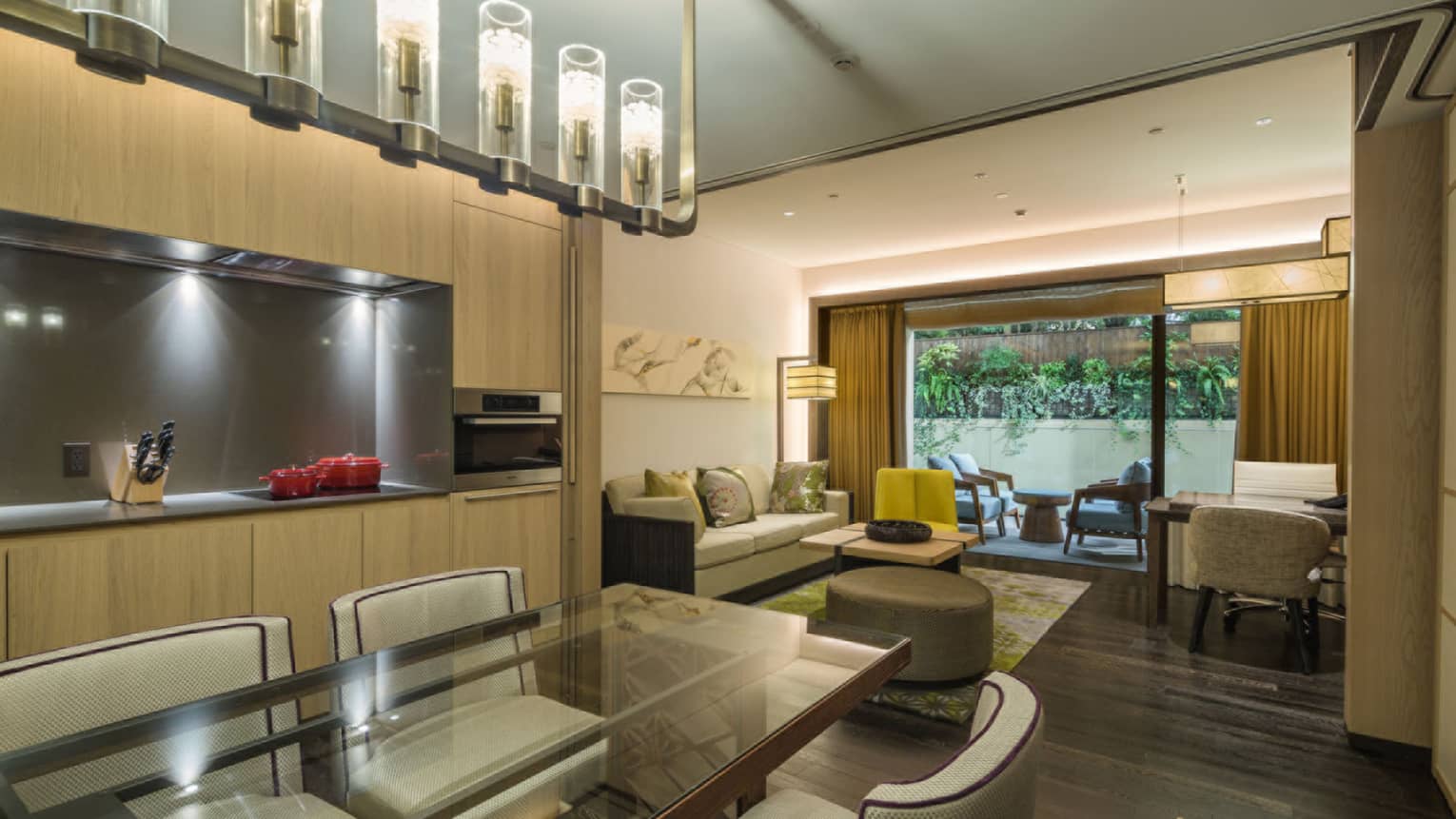 Luxury residential suite with dining table, kitchenette, sofa, desk and chairs, leading to a patio with outdoor seating and view of lush greenery