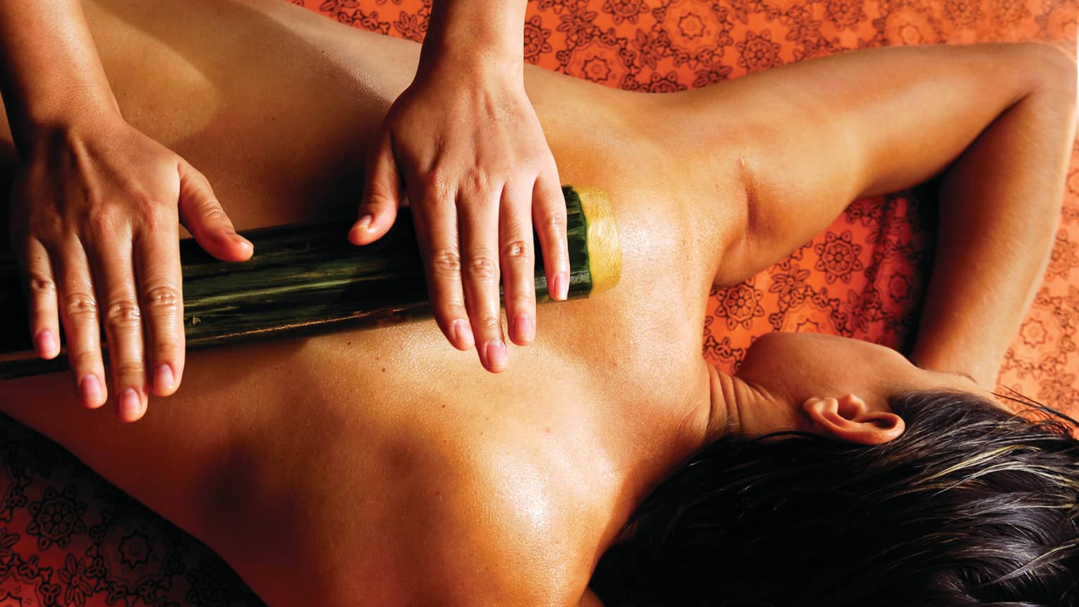 Spa staff rolls thick bamboo stick over woman's bare back, shoulders