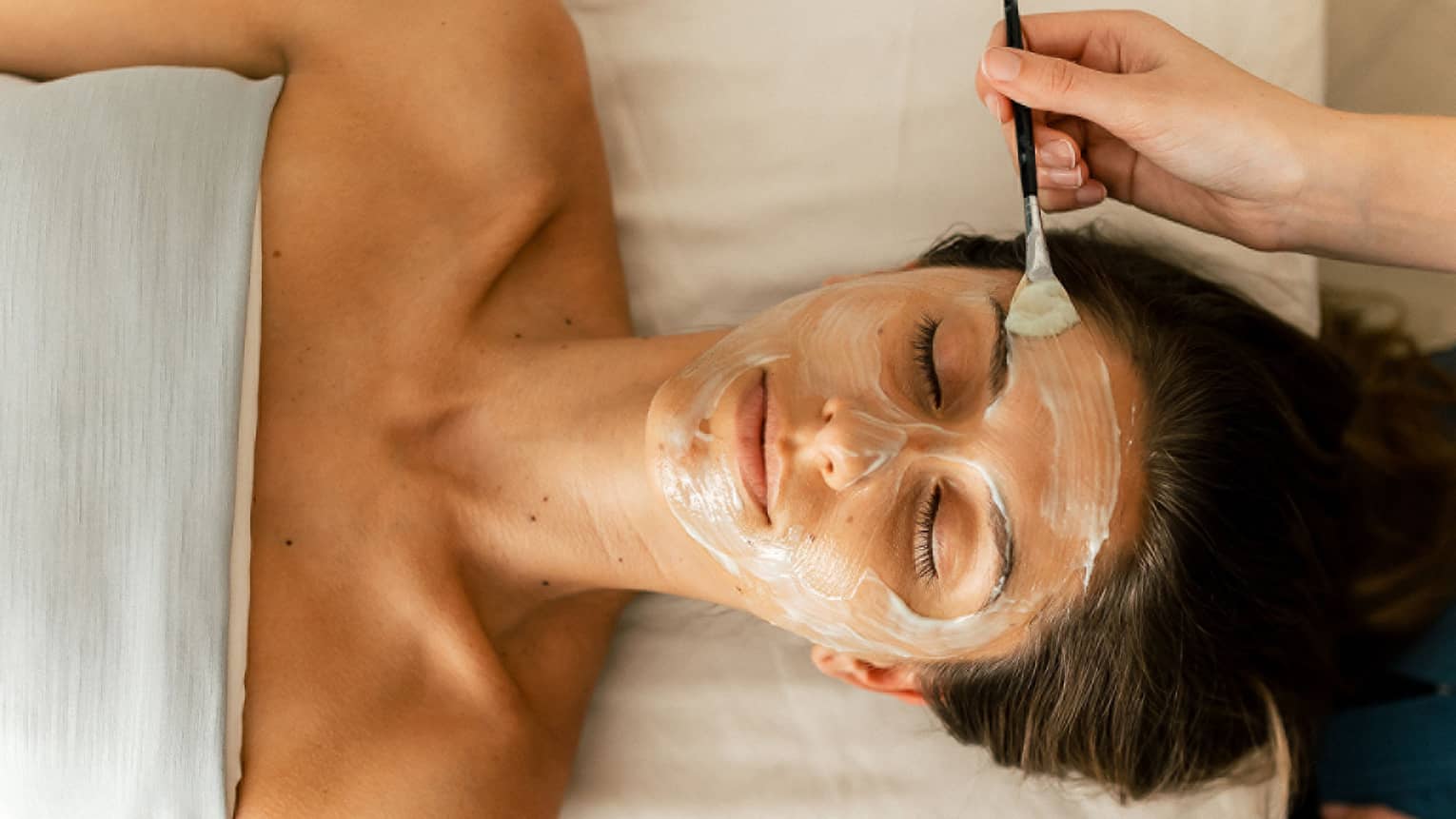 A person holding a small brush applies a white face mask treatment onto a woman's face