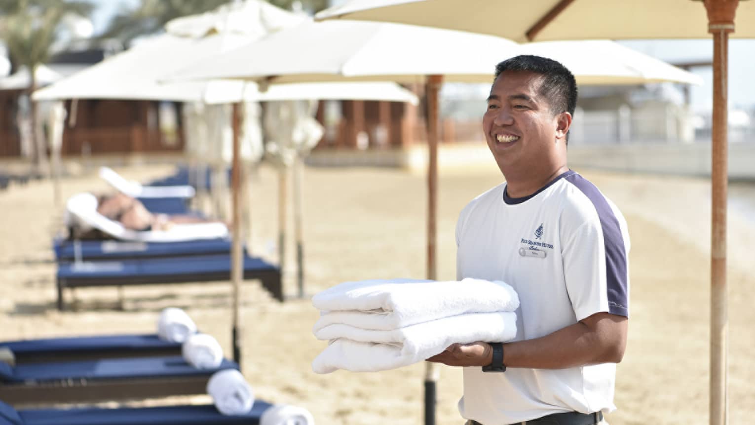 Beach attendant wearing white t-shirt and khaki pants smiles as he carries two fresh towels past navy-blue lounge chairs on the beach