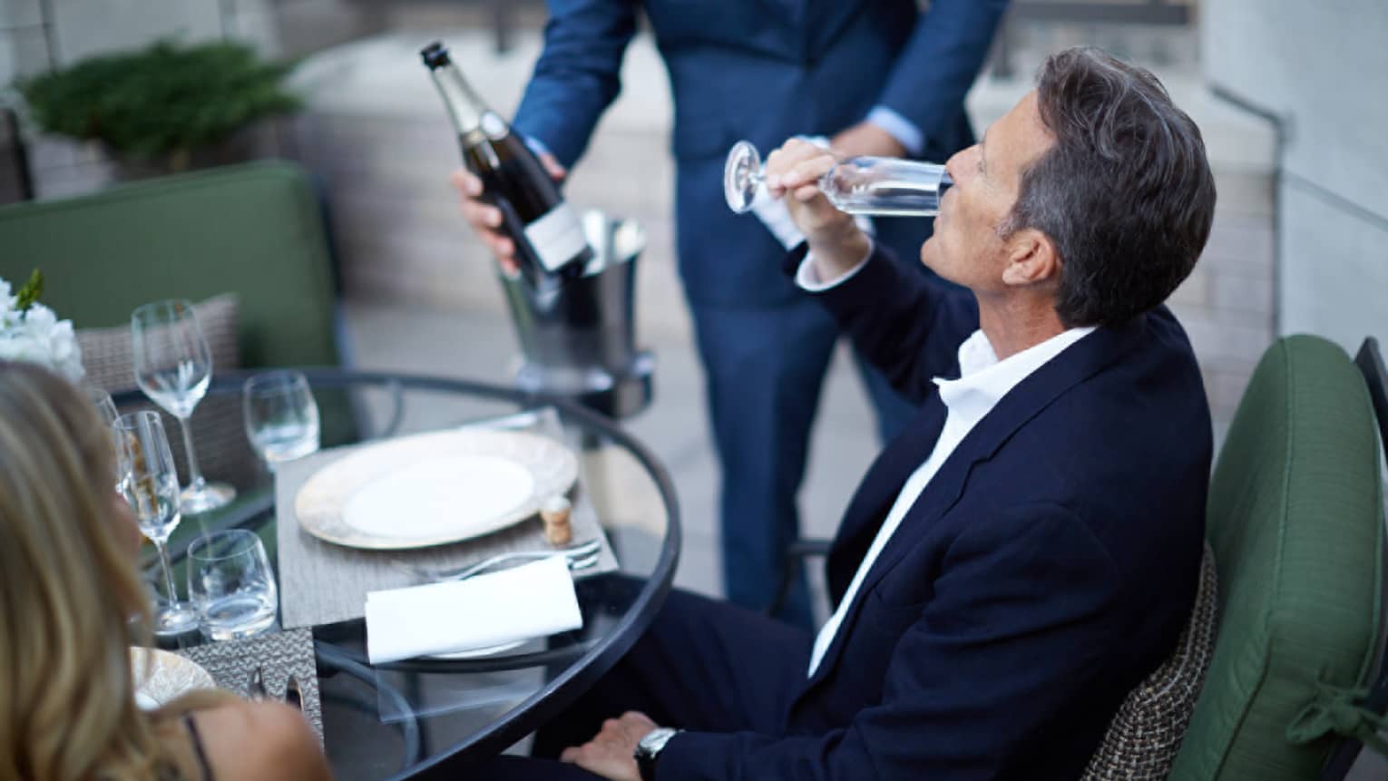 Patio dining table, man wearing suit sips from Champagne glass 