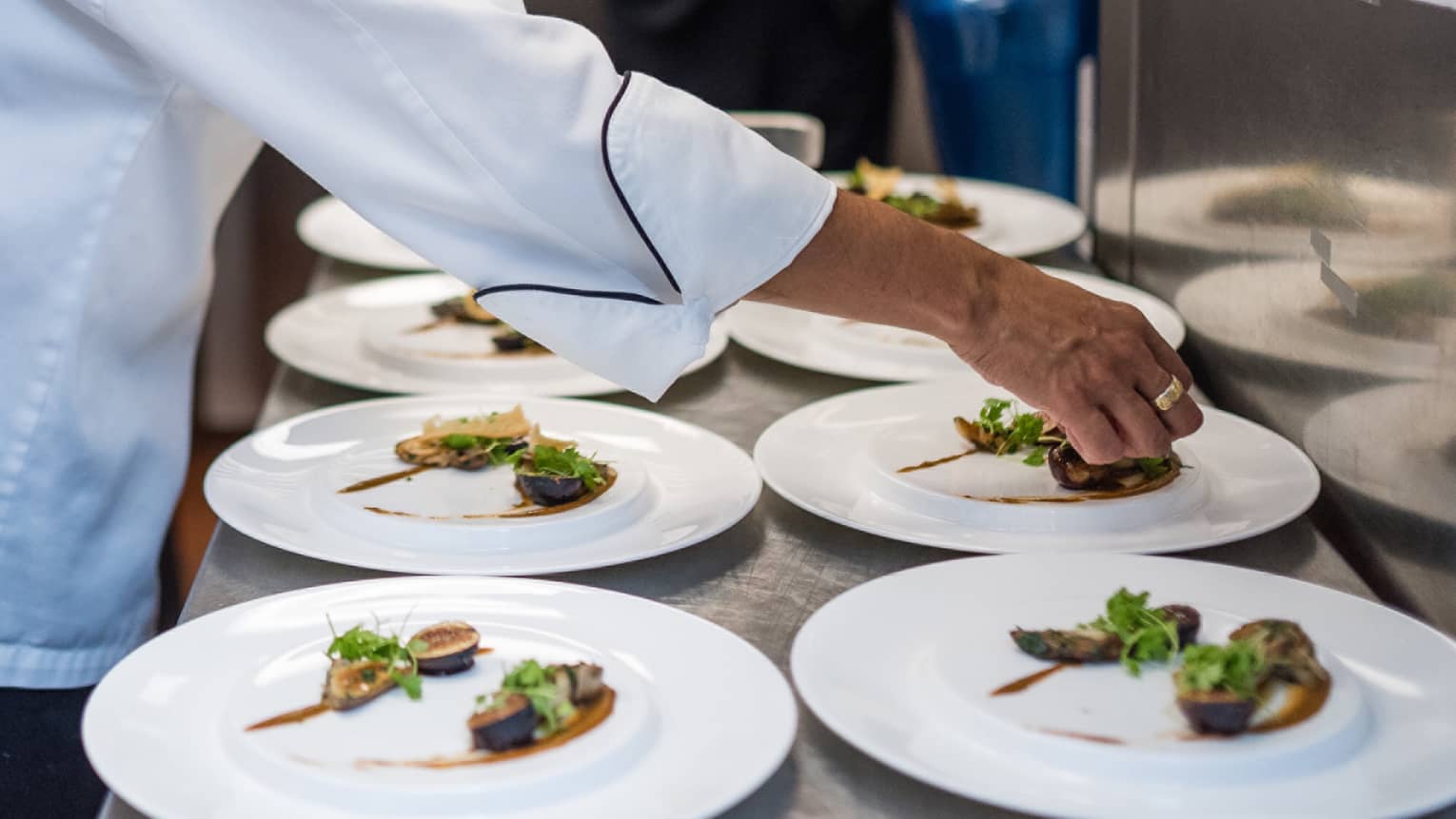 Chef plating figs and fresh greens on rows of white dinner plates