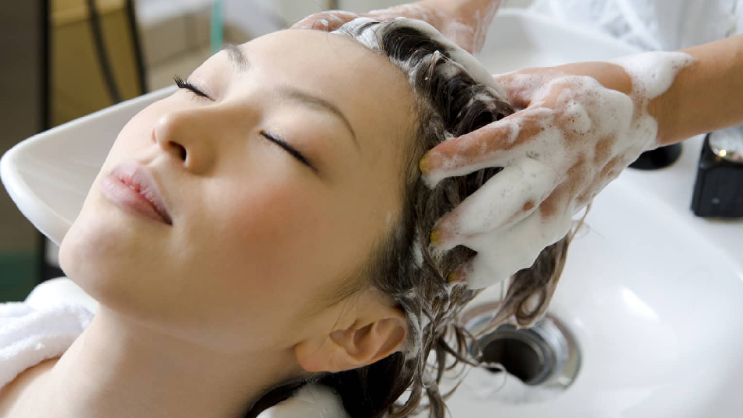 Woman closes eyes and leans back against sink as hands massage shampoo into her scalp