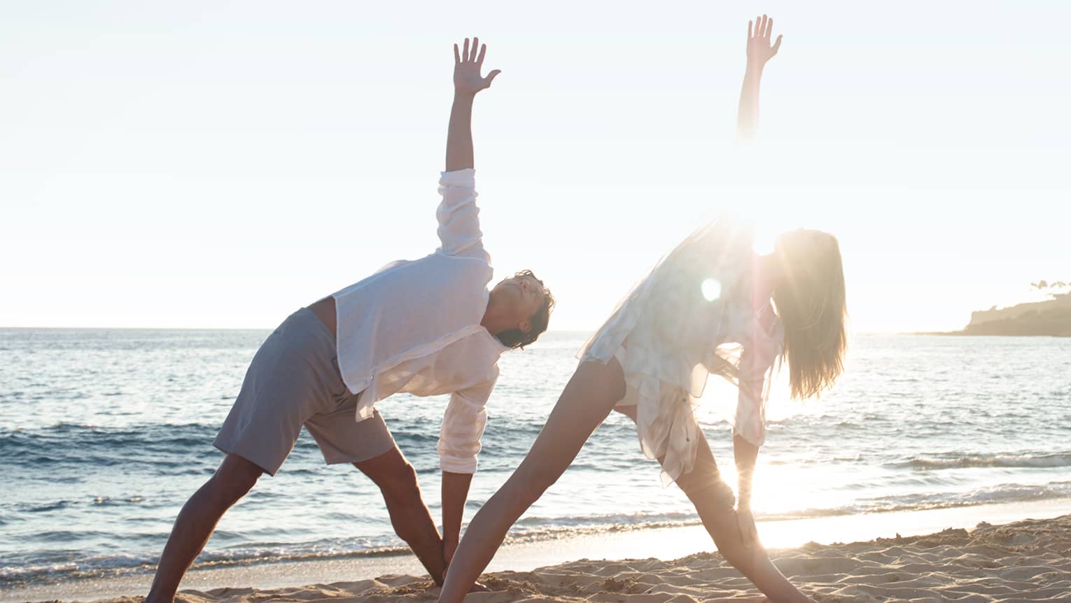 Two people in white beach shirts and shorts do a yoga pose with yogi at sunrise on beach