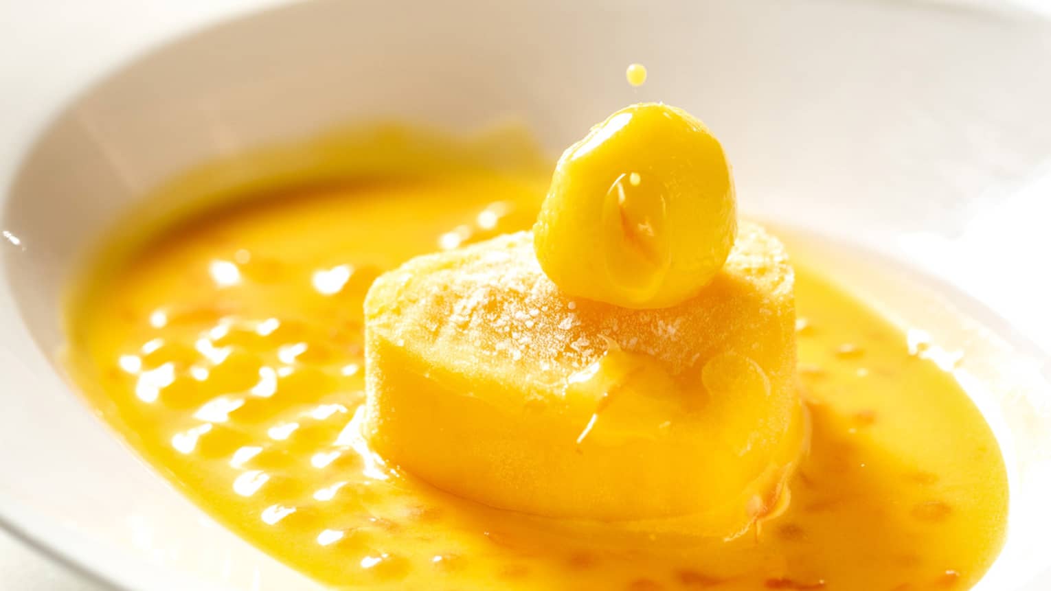 A white spoon drips yellow sauce onto a vibrant mango dessert served in a white dish
