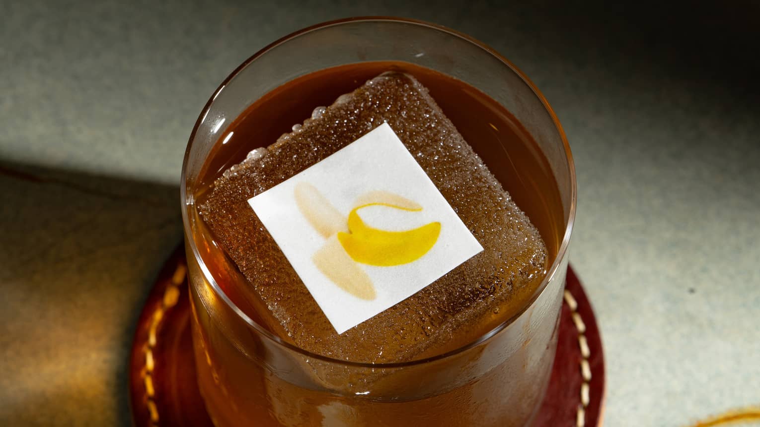 An amber-hued cocktail in a clear tumbler with a topping showing  an image of a banana