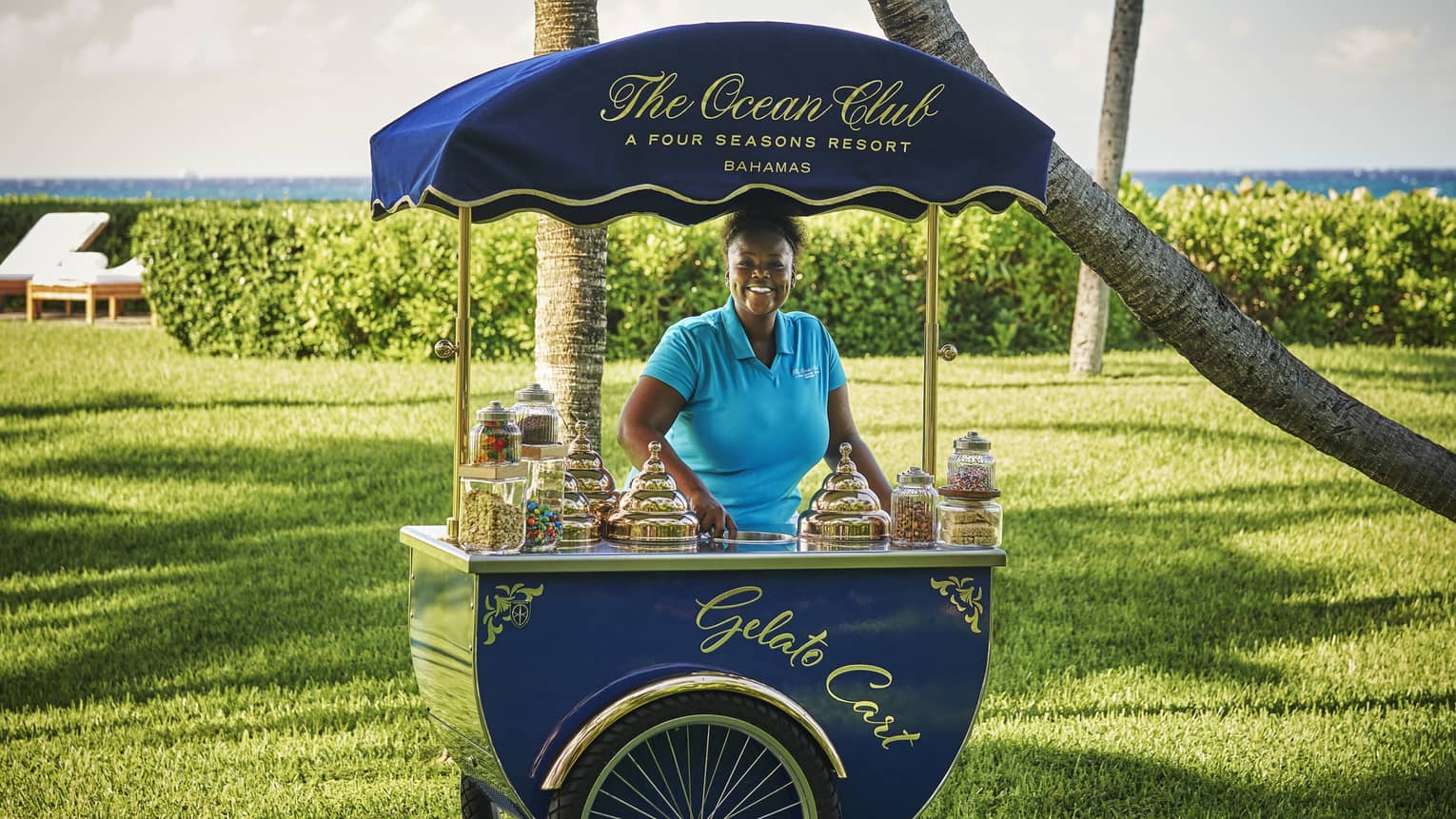 Smiling woman standing behind a blue gelato cart by The Ocean Club, Four Seasons Resort Bahamas, with a green lawn and ocean view in the background