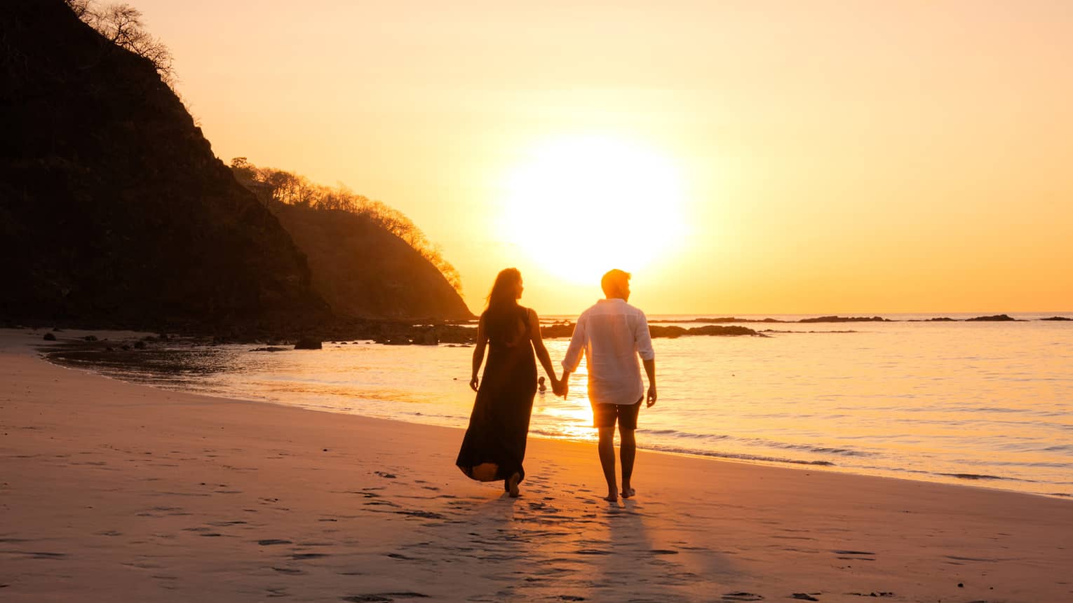 Couple walks hand in hand along the beach, silhouetted by the setting sun