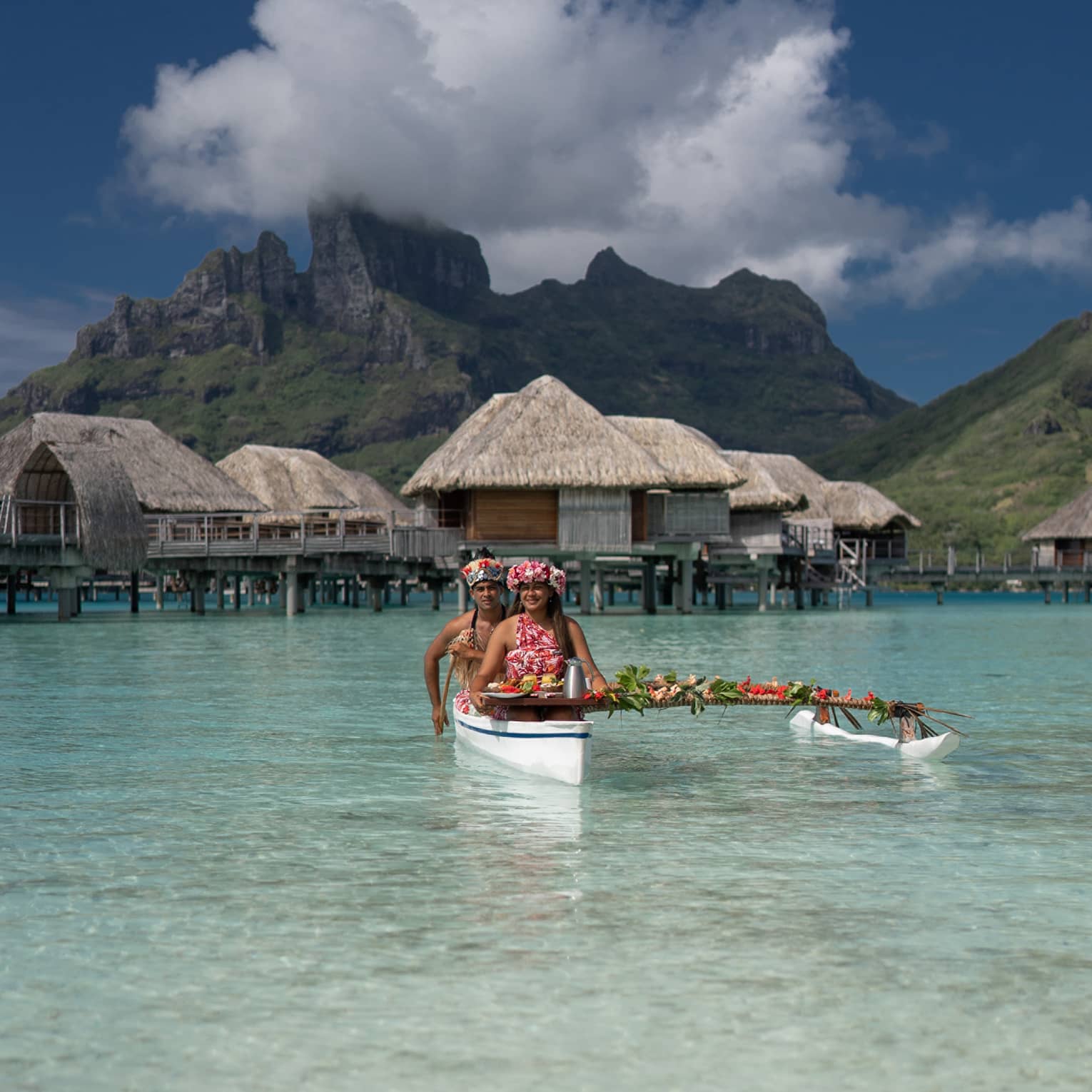 Two vibrantly dressed people in a canoe proffer a tray with colourful food attached to a pole draped with leaves and flowers.