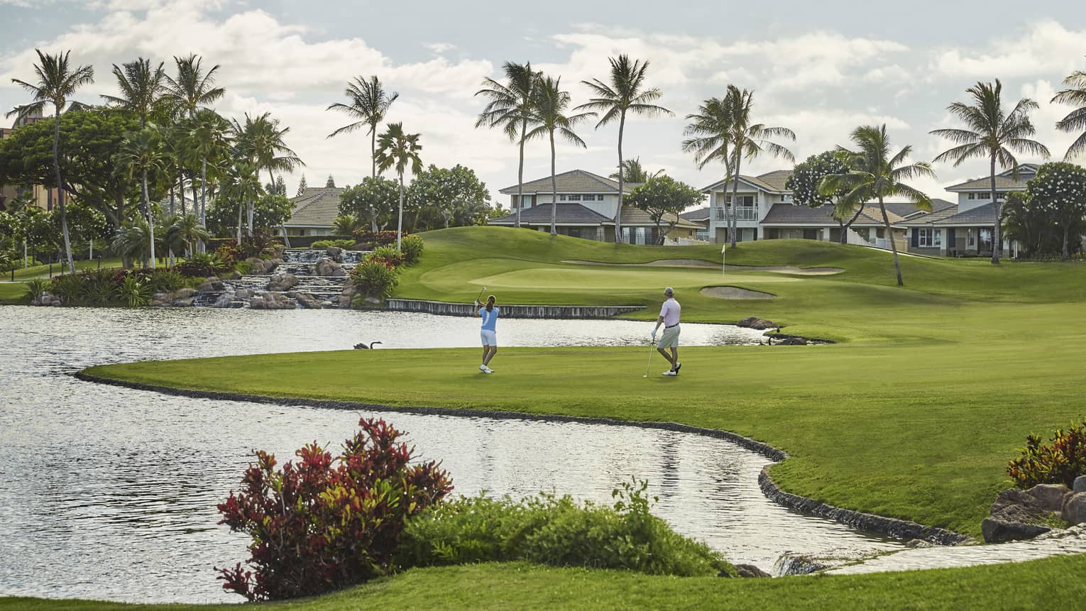 Two golfers on green grass near pond, palm trees in the distance