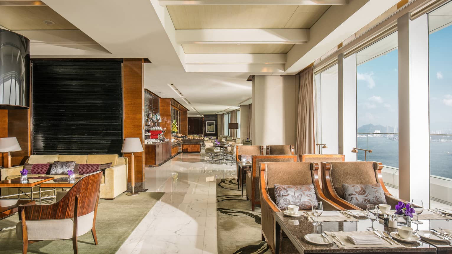 Hotel lounge with modern wooden chairs and set dining tables lined up along tall windows with water view