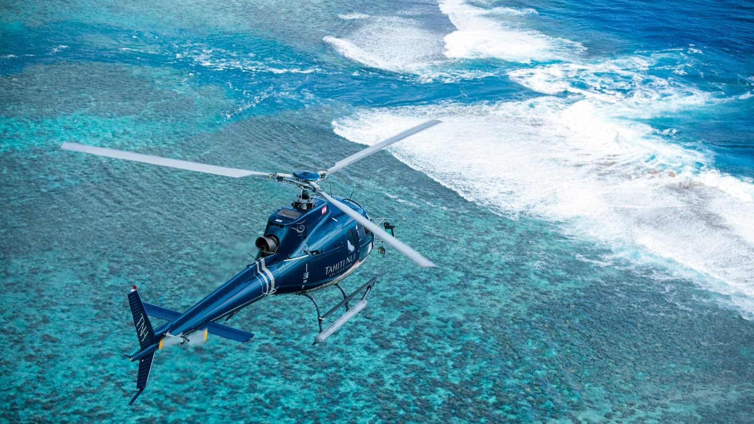 Bird’s-eye view of a helicopter flying over crystal-clear teal water with frothy waves breaking over a sandbar.