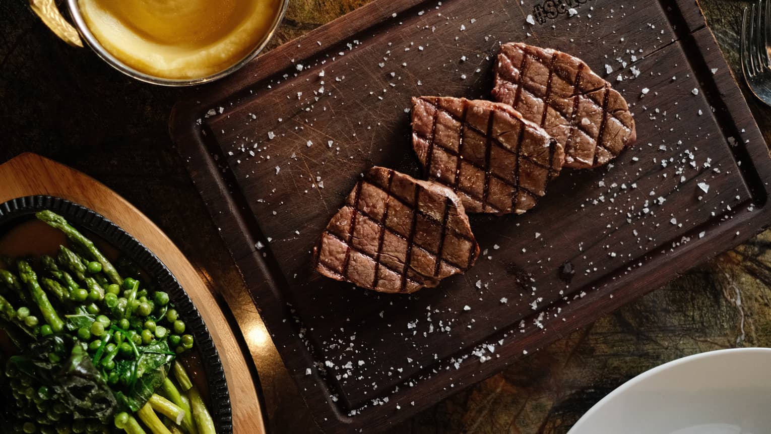 Three pieces of grilled steak on a dark wood cutting board, sprinkled with salt