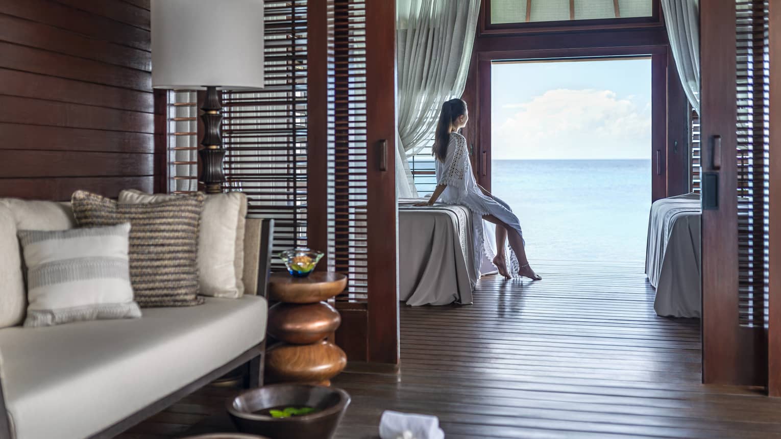 A woman leans on a massage bed on the far side of a spa treatment room and looks out through a large doorway onto the vast blue ocean