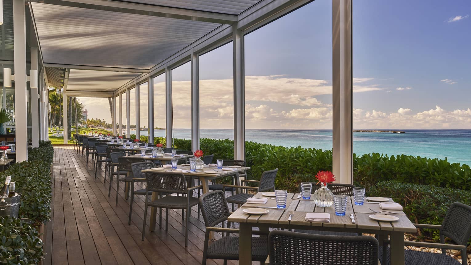 An outdoor eating area with square wood tables next to plants and the ocean.