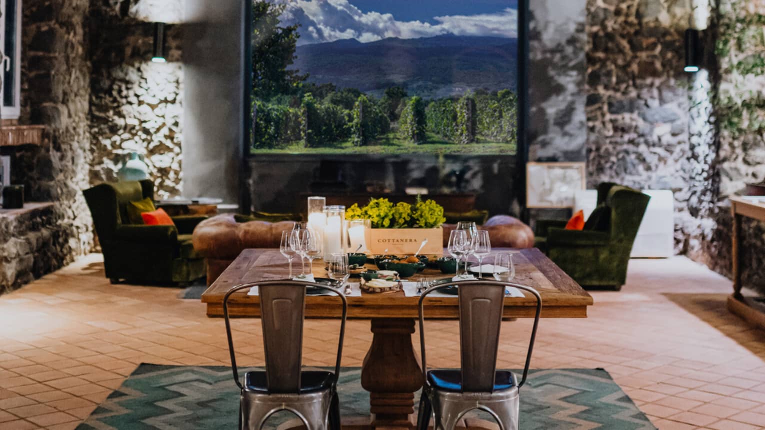 A wooden dining table set with floral arrangements and glassware,  in a room with a window overlooking mountains and a vineyard