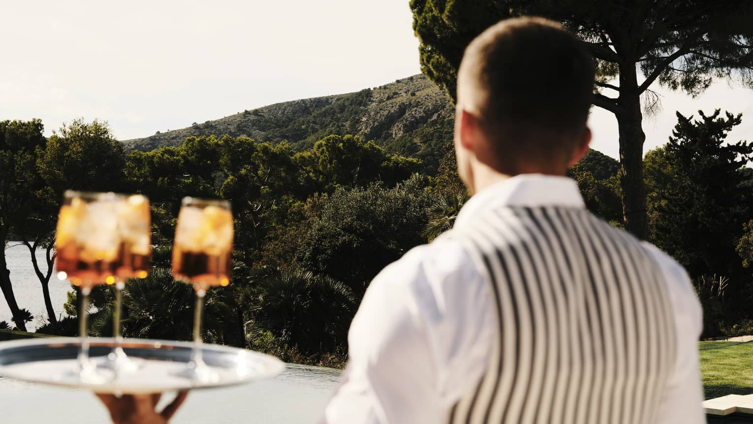 A server in a striped vest carries three cocktails on a silver tray by a pool set against a backdrop of lush, forested hills.
