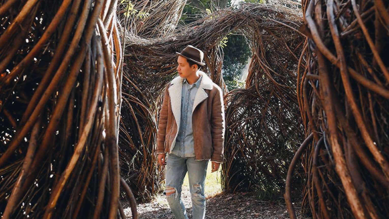 Man in shearling coat, fedora hat walks through twisted branches, trees
