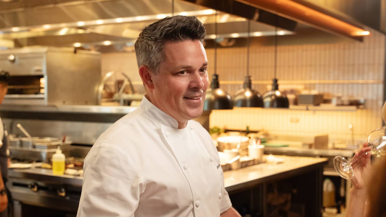 Chef standing beside a dining table in the kitchen talking to guests.
