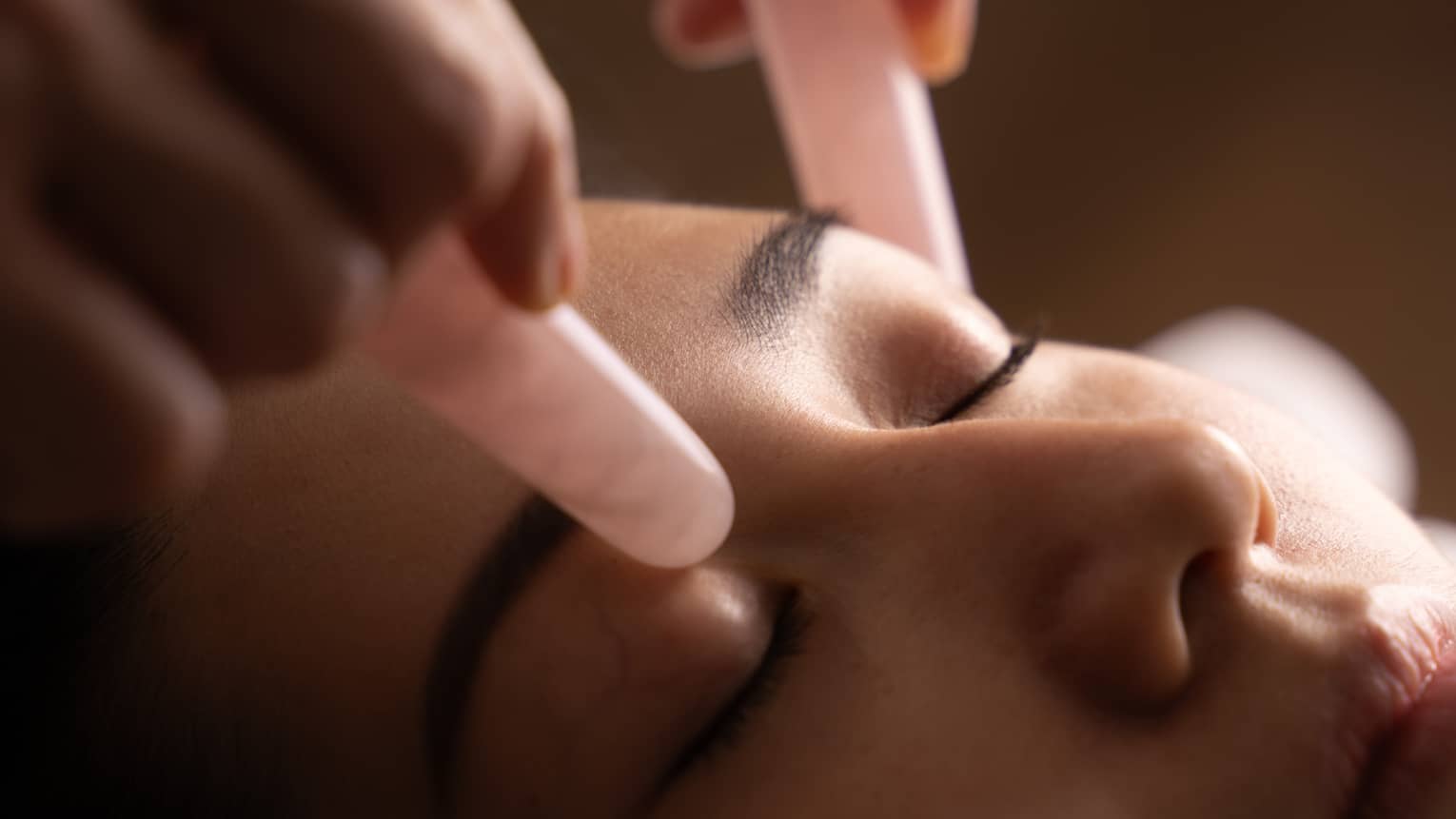 Hands hold pair of pink jade stones against woman’s eyelids during facial