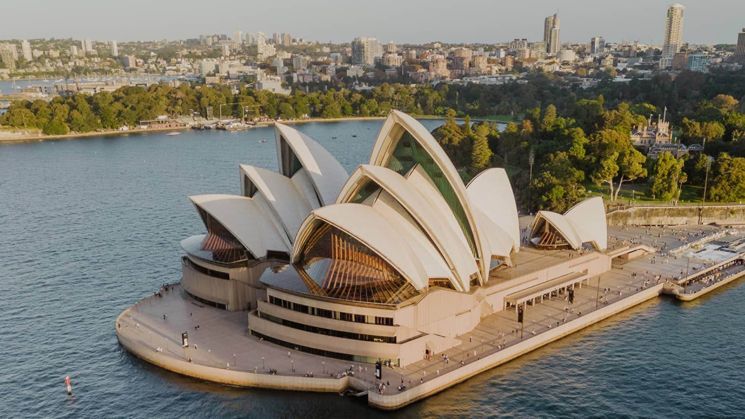 Aerial view of the Sydney Opera House, surrounded by deep blue harbour waters, with trees and city buildings beyond.