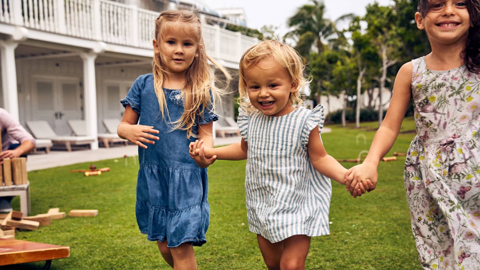 Three young girls holding hands as they play on grass next to a large white building.