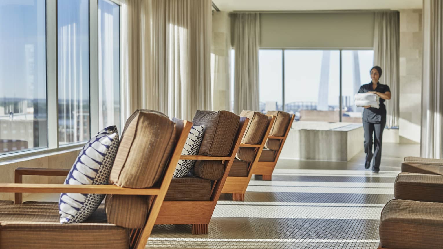 Spa attendant holding two folded white towels walks down sunny hall past lounge chairs facing windows