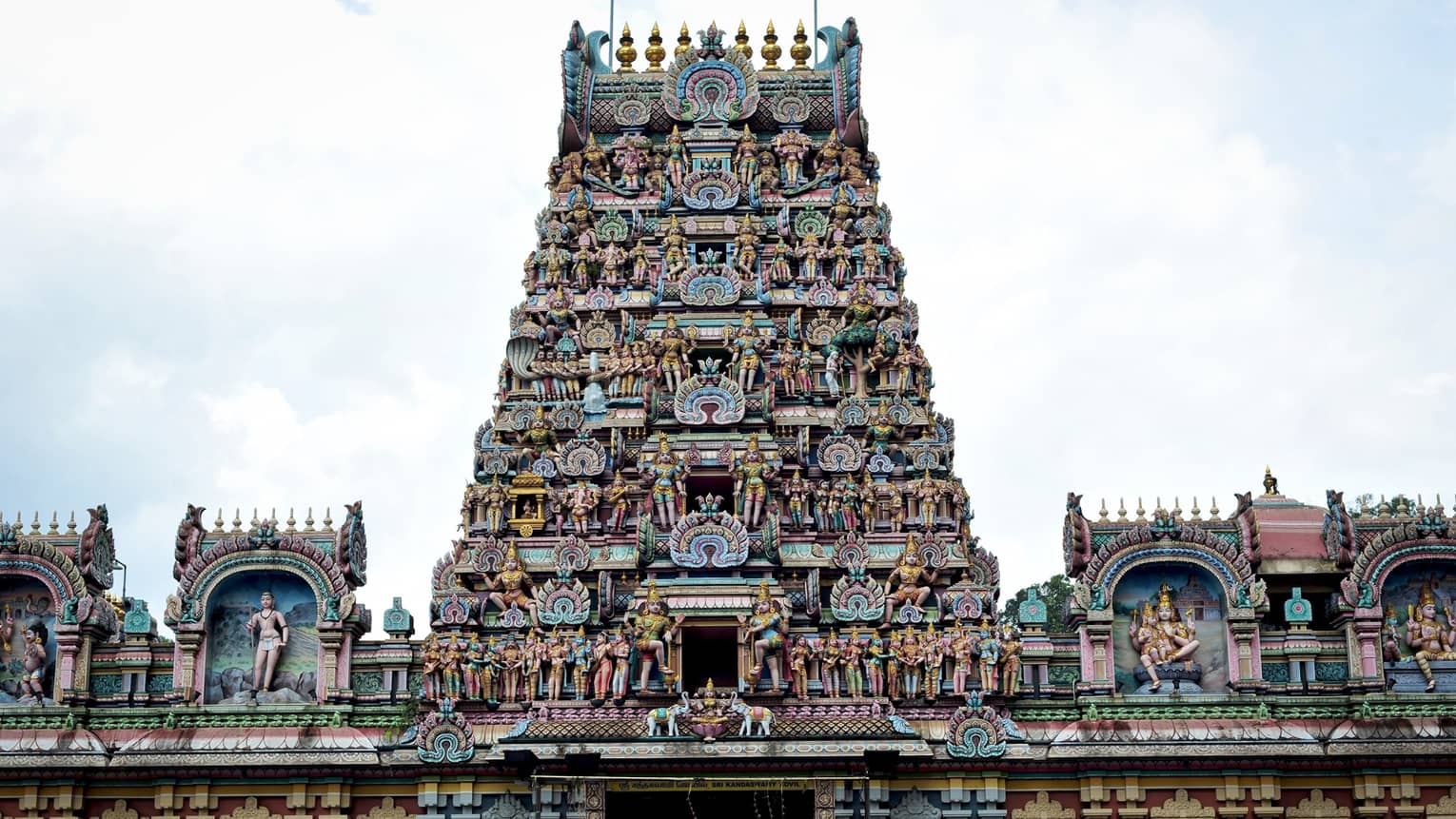 View from below of a temple adorned with intricate statues, topped by a colourful pyramidal structure