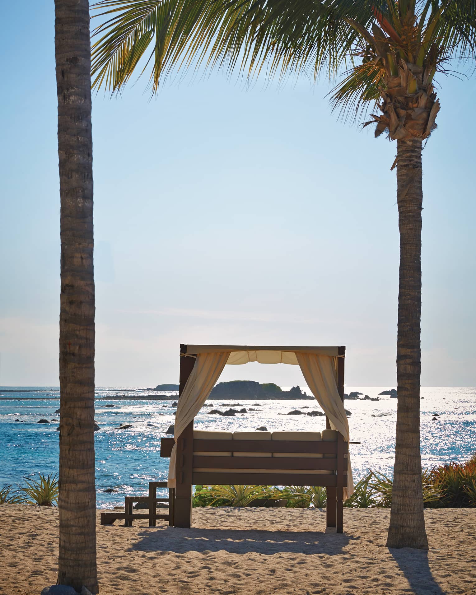 Steps lead up to a curtained daybed flanked by palm trees overlooking rocky islands in a sea of glimmering turquoise.