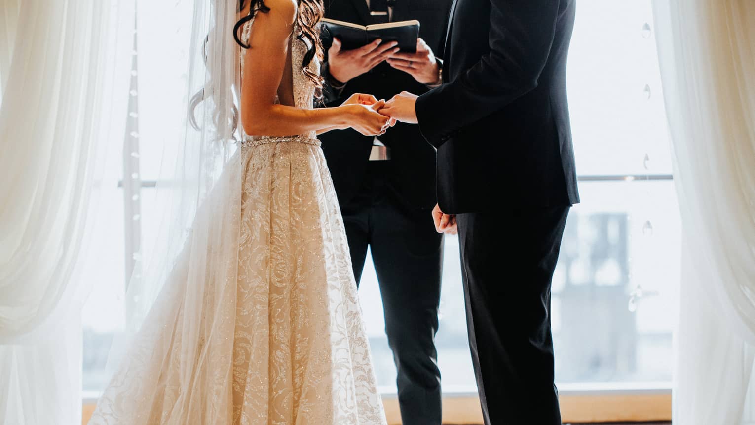 Bride and groom hold hands as wedding officiant reads from book by sunny window