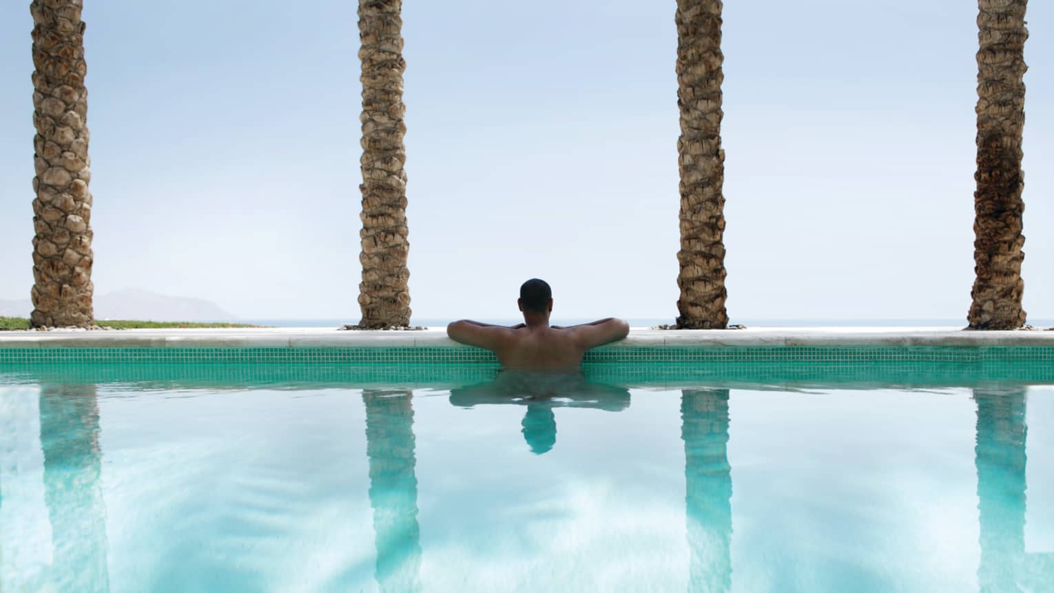 Back view of man resting arms on outdoor swimming pool ledge lined with palm tree trunks