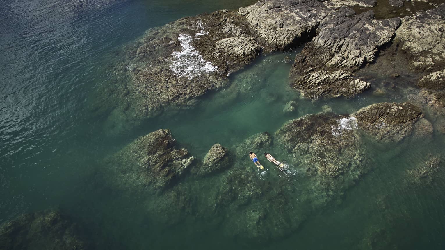 Aerial view of two people snorkelling by large rocks in the ocean 