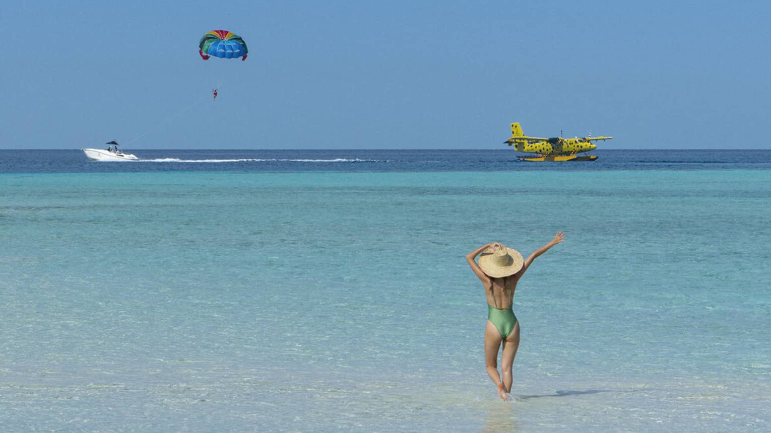 A woman walking on the beach watches a seaplane and a parasailor in the distance. 