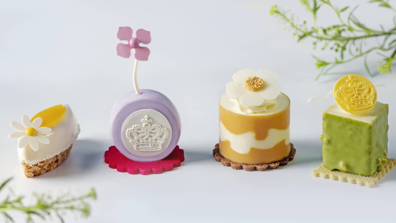 On a white table sit four small, pastel-coloured cakes in various shapes on cookie bases and topped with floral accents.