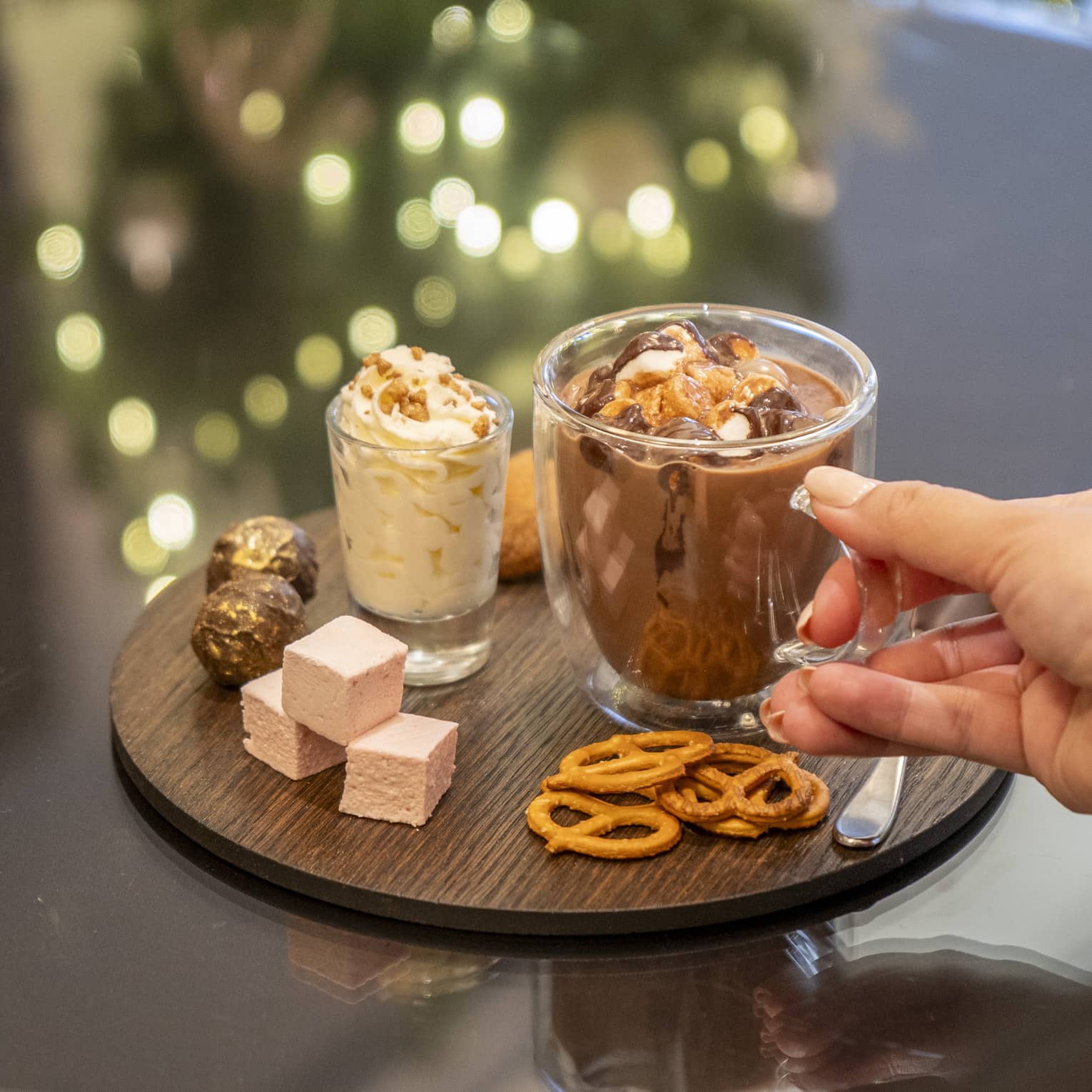 Round wooden platter with a glass mug of hot chocolate, small glass of whipped cream, thre pink marshmallow squares, pretzels and assorted chocolates