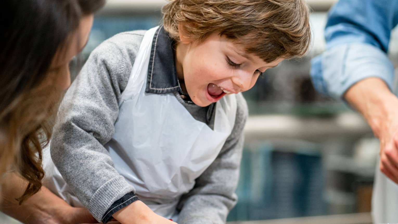 Aided by an adult, a child eagerly squeezes chocolate sauce out of a bottle onto a marble counter behind large cocoa beans. 