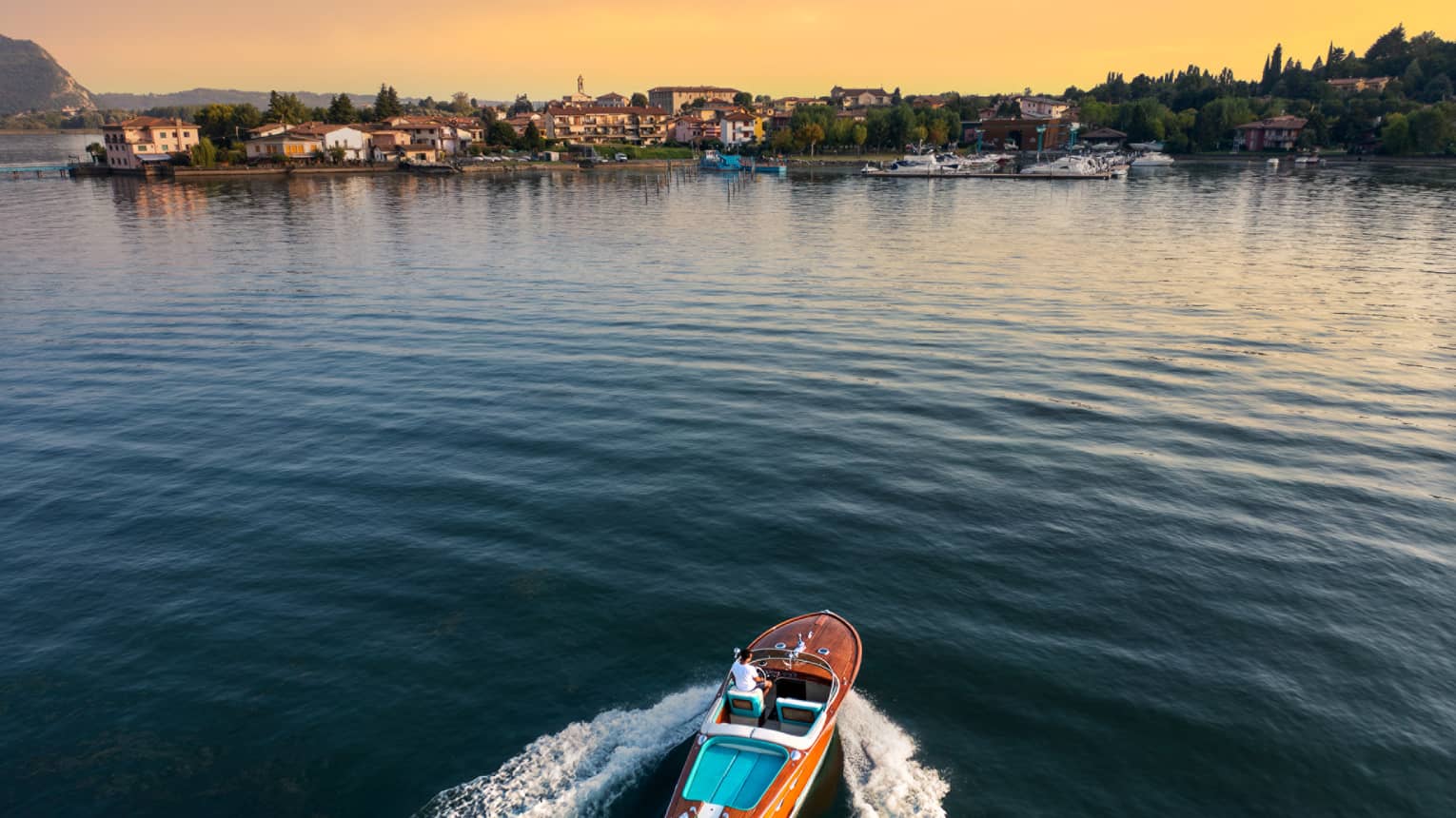 Person driving a wooden speedboat across a lake towards shore at sunset